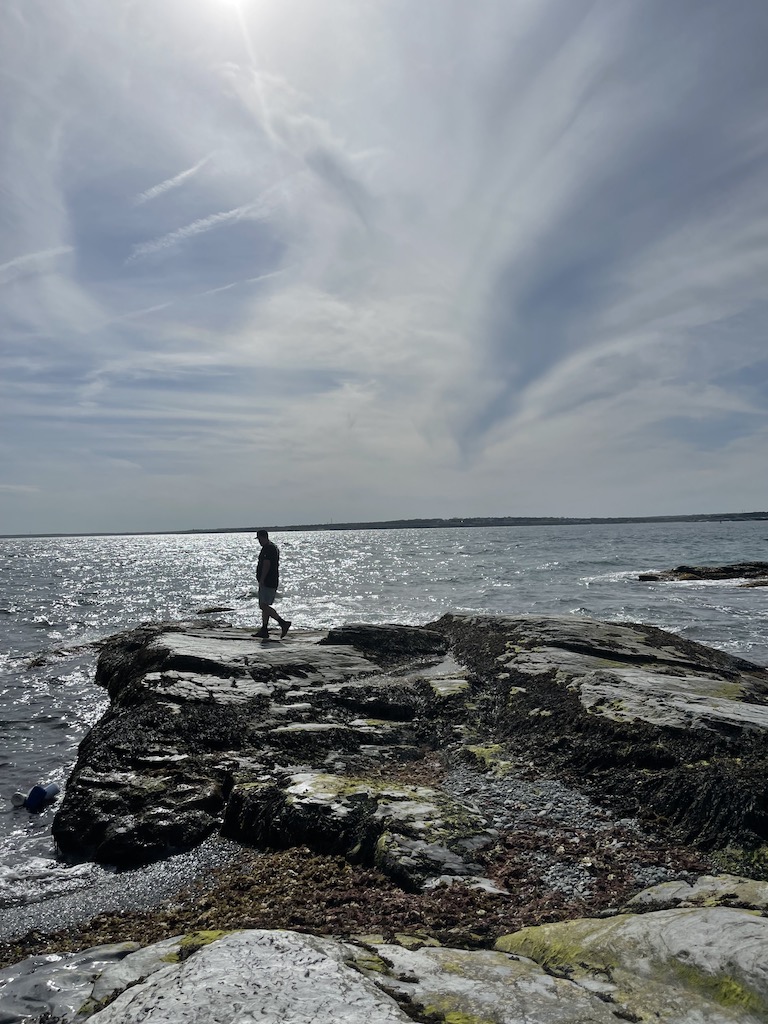 Silhouette of man walking on rocky shoreline of Atlantic Ocean on partly cloudy day. Brenton Point State Park, Newport, Rhode Island.