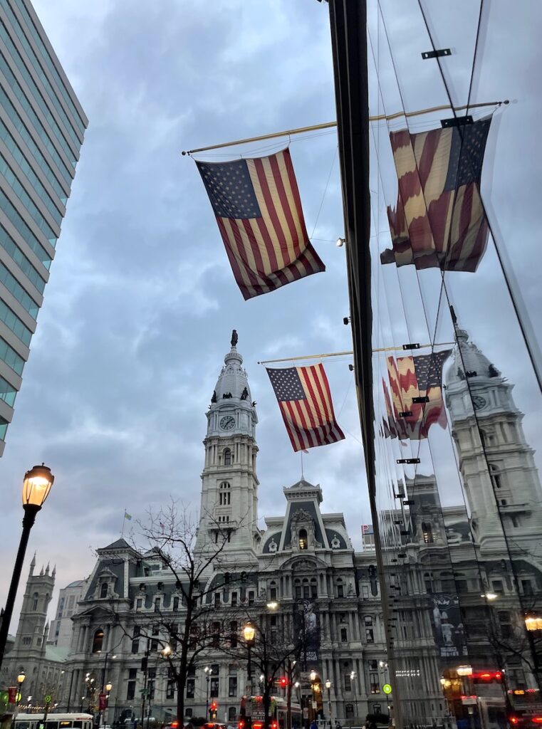 American flags reflecting on windows of skyscraper building with large ornate government building in background on grey evening. Philadelphia, Pennsylvania.