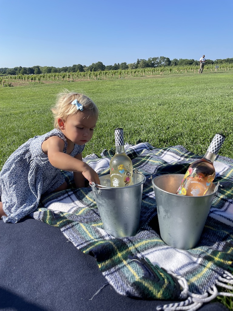 Toddler on picnic blanket reaching into ice bucket of holding bottle of wine, next to it sits another bucket of ice with bottle of wine, in background is grassy field leading up to grapes growing in a vineyard. The Wine Stand at Wolffer Estate Vineyard, Sagaponack, New York, The Hamptons.