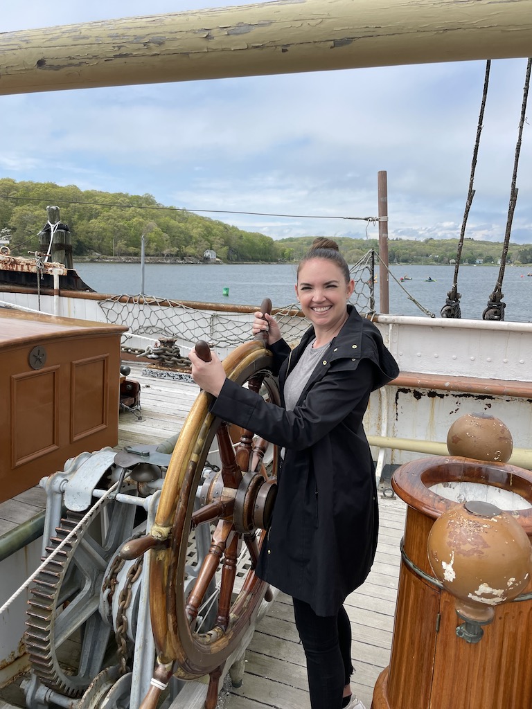 Woman pretending to steer historic wooden sailboat with huge wooden steering wheel with Mystic River and green hills in background on sunny day. Mystic Seaport Museum, Mystic, Connecticut, New England.