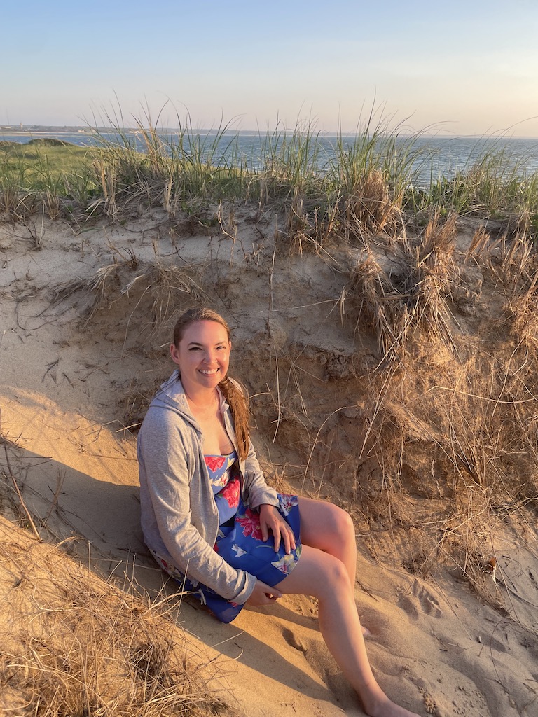 Woman sitting on beach sand dune with Atlantic Ocean in background during sunset. First Encounter Beach, East Ham, Cape Cod, Massachusetts, New England.