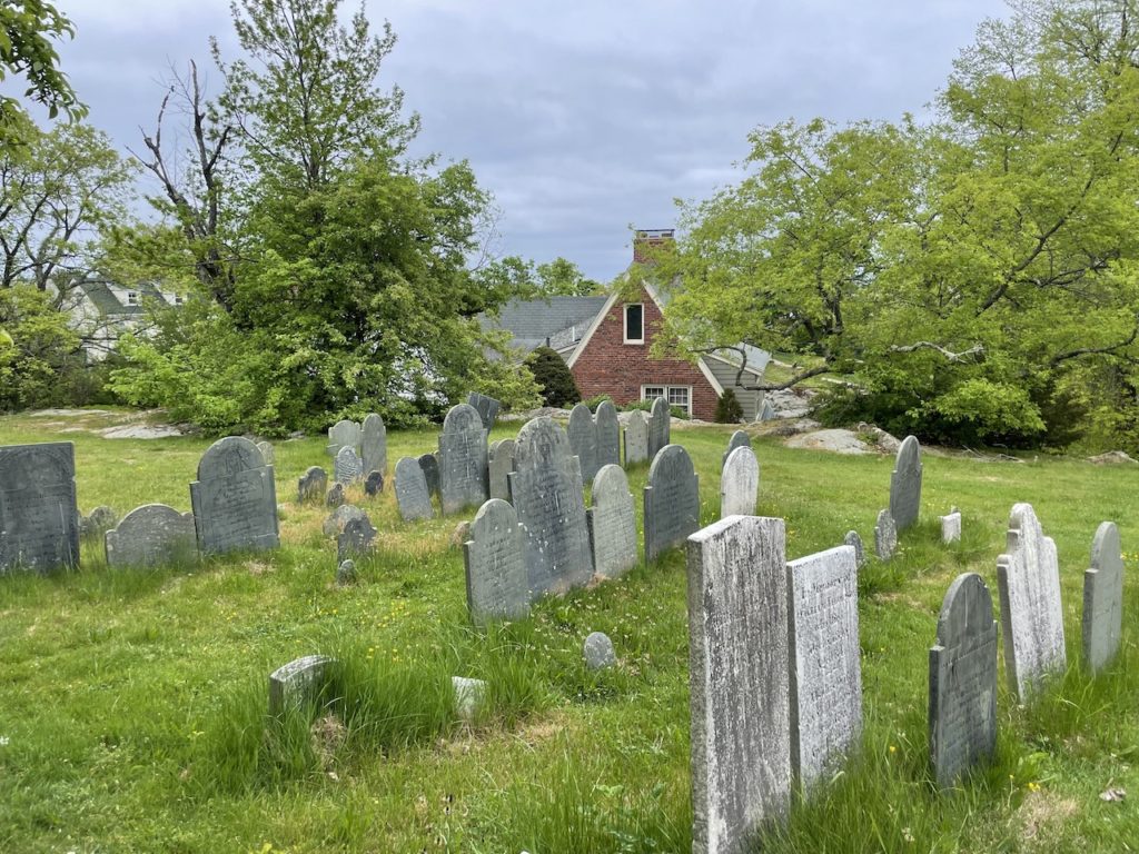 Old stone tombstones on grassy hill with green trees and historic brick house in background on gloomy day. Old Burial Hill, Marbleville, Massachusetts, near Salem, New England.