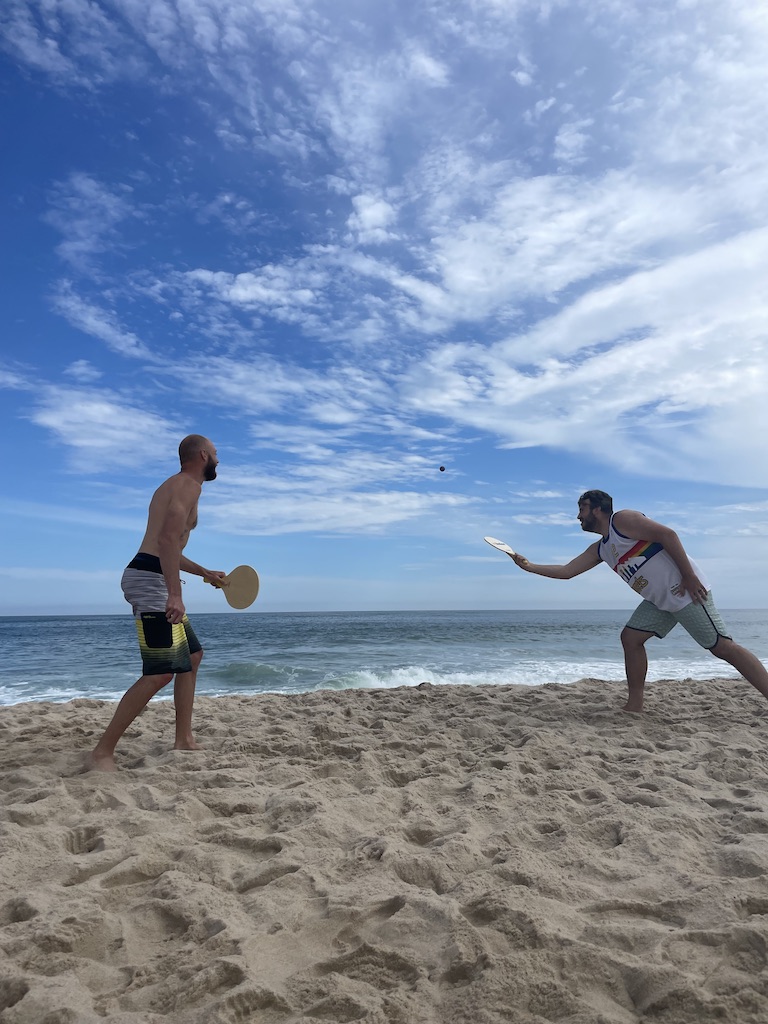Two men on white sand beach with Atlantic Ocean in background planning Kadima, a game with two wooden paddles and small rubber ball. Sagg Main Beach, Sagaponack, New York, The Hamptons.