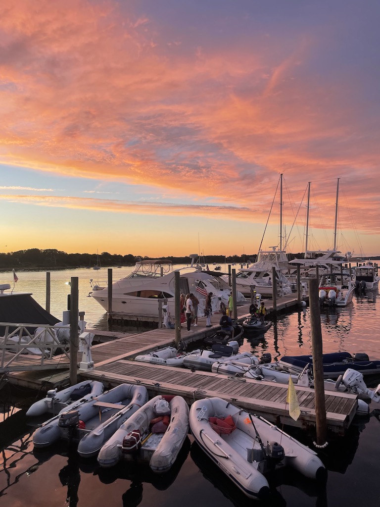 Boats of all sizes docked on wooden dock over calm harbor water with colorful sunset in background. Sag Harbor Yacht Yard, Sag Harbor, New York, The Hamptons.