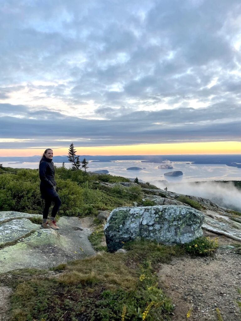Woman on mountaintop overlook looking down on the Atlantic Ocean during foggy sunrise. Cadillac Mountain sunrise, Acadia National Park, Maine, New England, United States.
