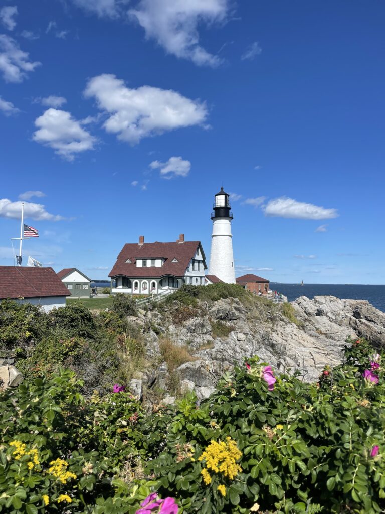 White lighthouse on edge of rocky shoreline with ocean in background on sunny day. Portland Head Light, Portland, Maine.