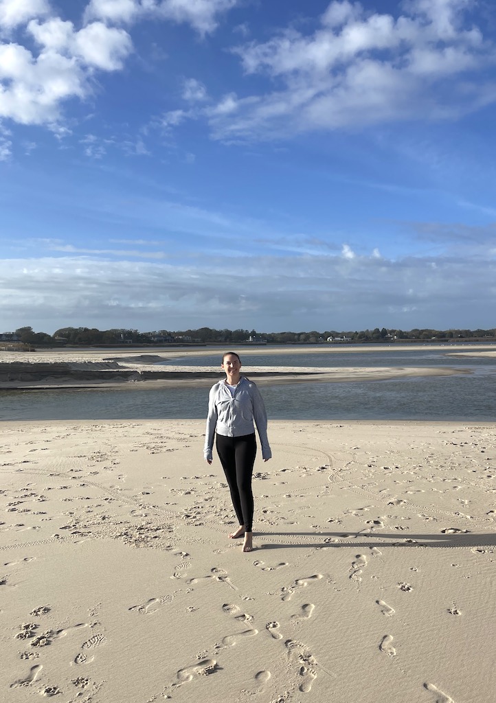 Barefoot woman in light jacket and pants on white sand beach with ocean inlet behind her on sunny fall day. Sagg Main Beach, Sagaponack, New York, The Hamptons.