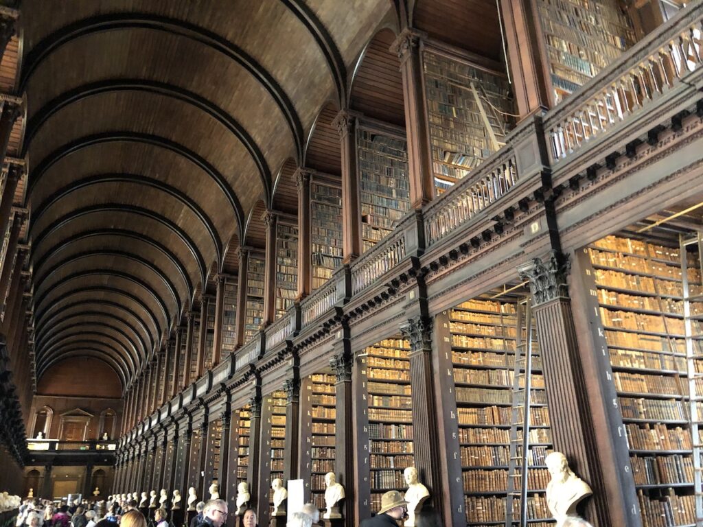 Massive, two story library with arched ceiling, everything is dark wood with marble busts lining walkway. Books are organized on shelves extending from the floor up to the second story of building. Long Room, Old Library, Trinity Library, Trinity College, Dublin, Ireland.