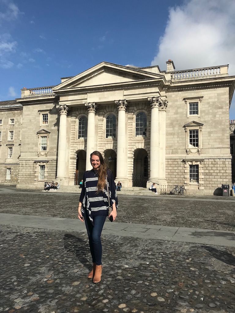 Woman walking on cobblestone street in front of ornate marble 3-story building with 4 columns. Trinity College, Dublin, Ireland.