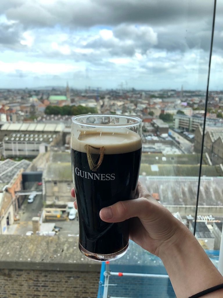 Pint of Guinness stout beer being held in front of large glass window with sprawling city views of downtown Dublin on grey day. Guinness Storehouse Gravity Bar, Dublin, Ireland.
