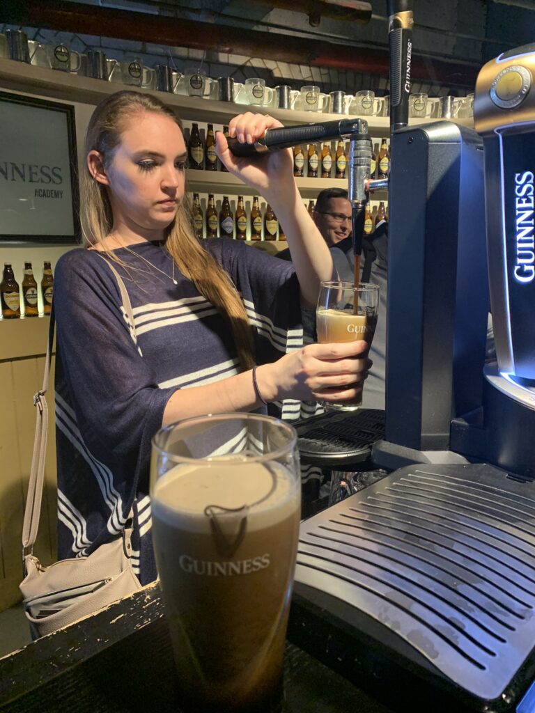 Woman pouring stout beer in bar-like setting, another pint of Guinness beer sits in foreground. Guinness Storehouse Brewery, Dublin Ireland.