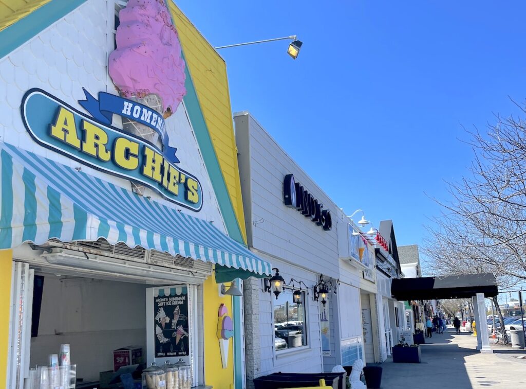 Colorful ice cream shop with pink/yellow/teal ice cream sign reading "Archies Homemade" colorful building sits along side row of storefronts on sunny summer day. Shopping on Rehoboth Avenue in Rehoboth Beach, Delaware.