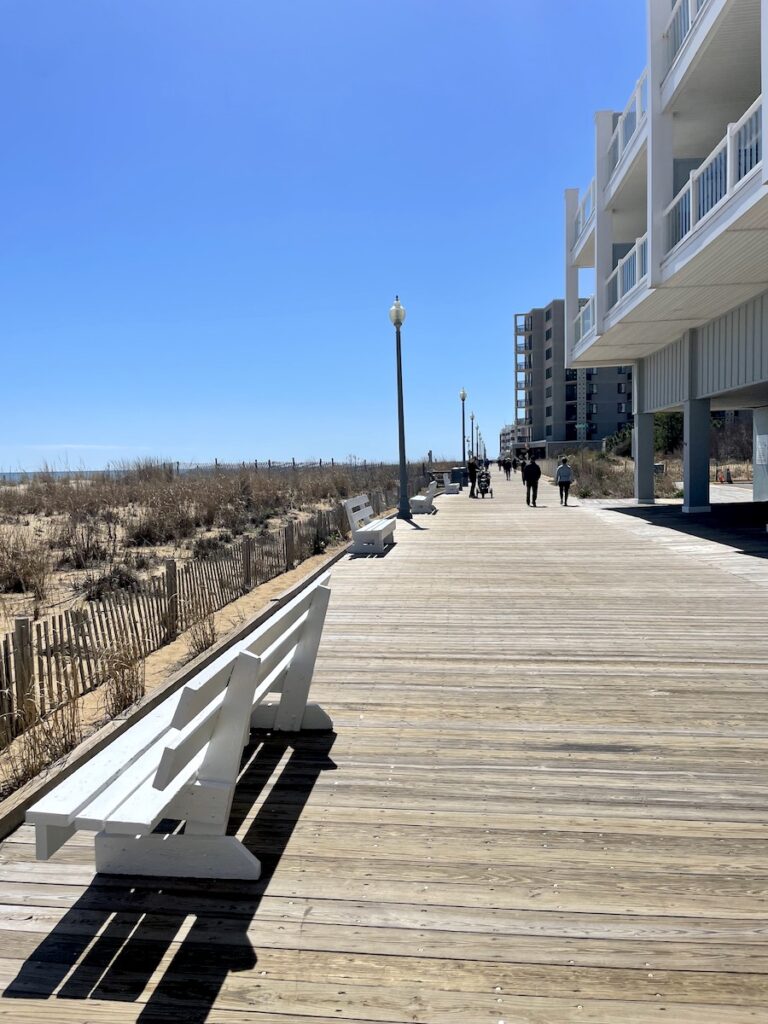 Wide wooden boardwalk with sand dunes and ocean to one side and beachfront businesses to the other on a sunny afternoon. Rehoboth Beach Boardwalk, Rehoboth Beach, Delaware.